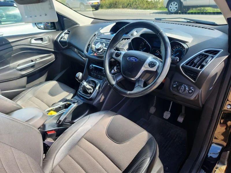 The front seats, steering wheel, and controls on a black Ford Kuga 2.0 TDCi Titanium, viewed from the right side.