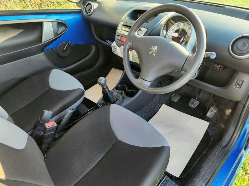 The front seats, steering wheel, and controls on a blue Peugeot 107 1.0 Access, viewed from the right side.