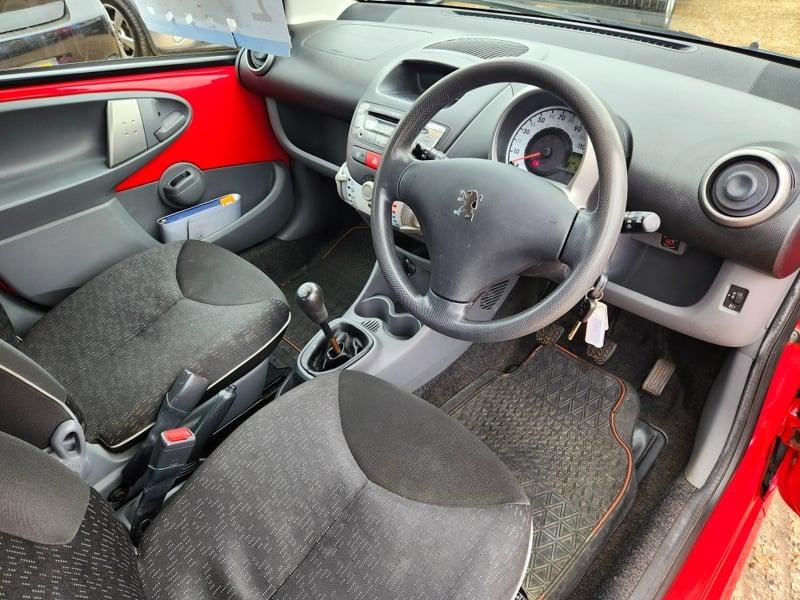 The front seats, steering wheel, and controls on a red Peugeot 107 Urban, viewed from the right side.
