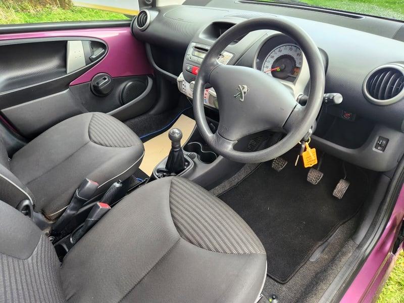 The front seats, steering wheel, and controls on a purple Peugeot 107 1.0 Access, viewed from the right side.