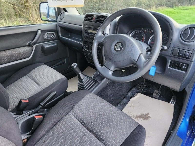 The front seats, steering wheel, and controls on a Baltic Blue Suzuki Jimny 1.3 VVT JLX, viewed from the right side.